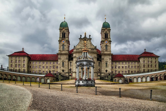 Обои картинки фото швейцария, айнзидельн, города, здания, дома, архитектура, замок, облака, башня, с, часами, скульптура, христианство, корона, арка